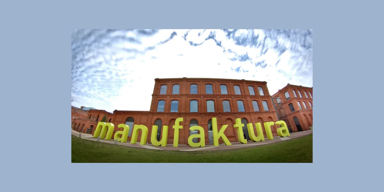 A wide-angle view of the Manufaktura complex in Lodz, Poland, with its distinctive red brick buildings and the large green "manufaktura" sign in the foreground.