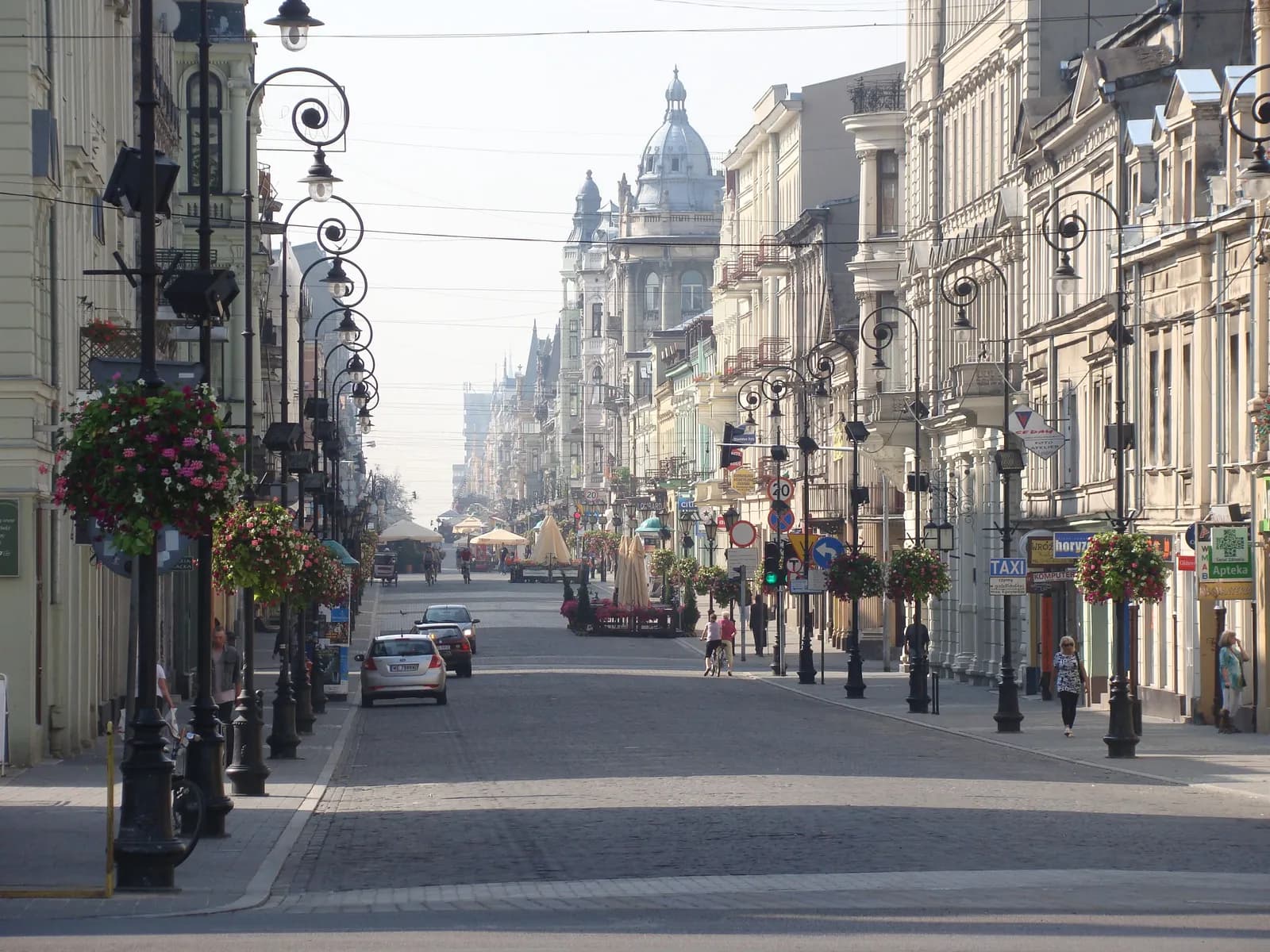 A long, historic city street lined with ornate lampposts holding hanging flower baskets, leading to distant buildings under a bright sky.