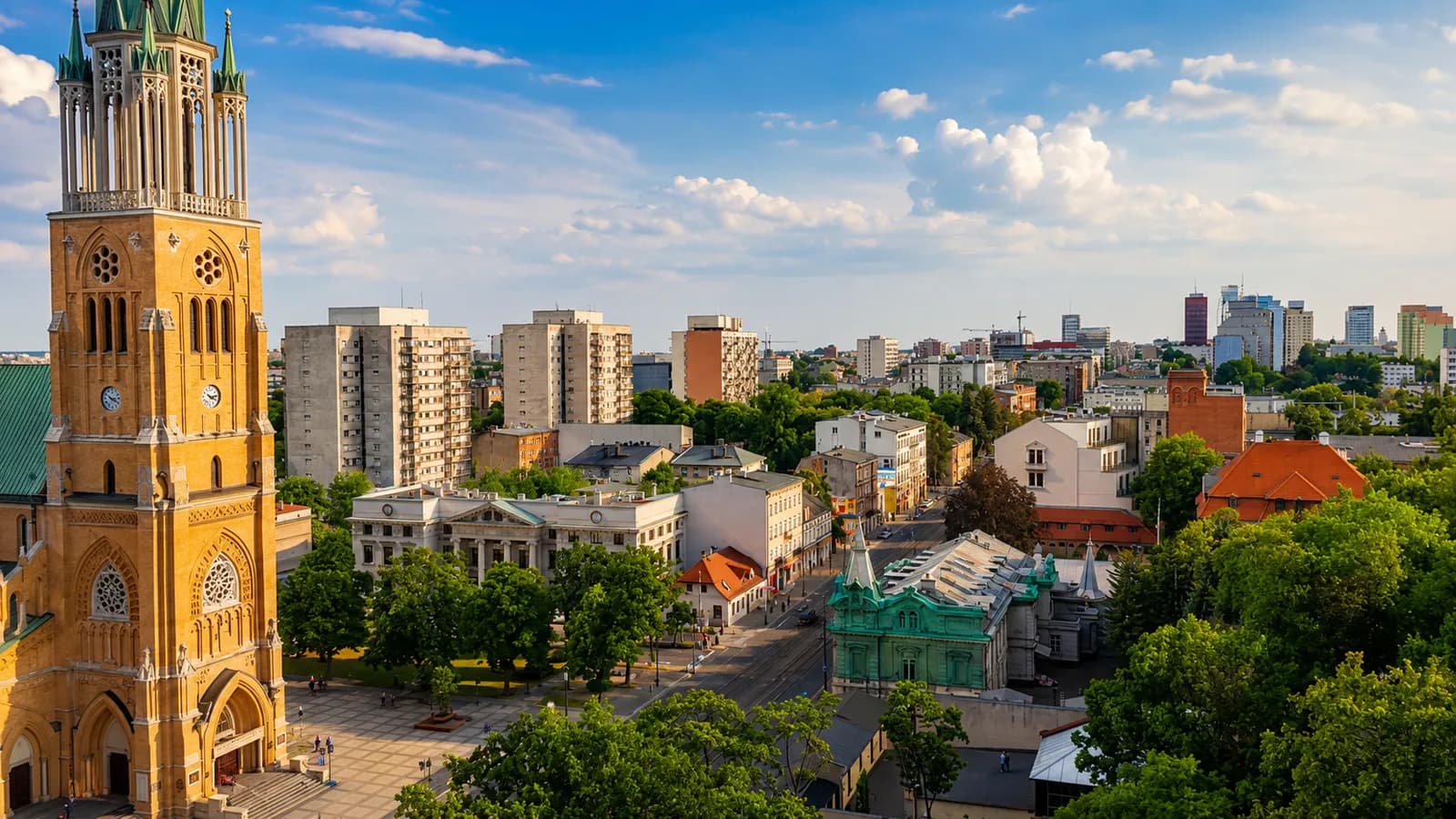 An aerial view of a European city with a large church tower in the foreground, surrounded by buildings and trees under a partly cloudy sky.